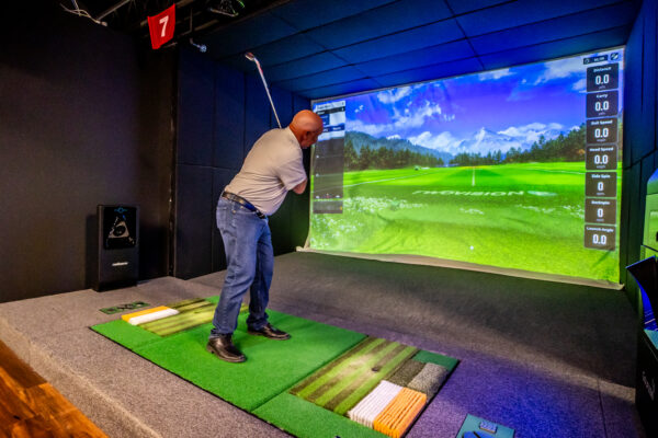 A man in a white polo and jeans swings a golf club in an indoor simulator bay. The large screen displays a high-definition image of a bright green golf course with mountains in the distance, while a digital sidebar shows data for distance, ball speed, and launch angle.