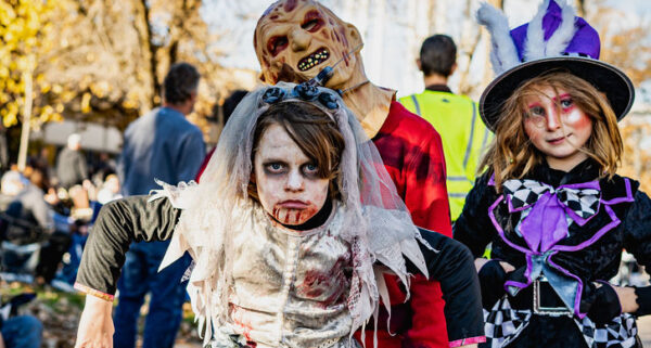 A young person in a bloody zombie bride costume looks directly at the camera, flanked by another person in a scary, monstrous mask and a person in a whimsical, Mad Hatter-style costume during a Halloween parade.