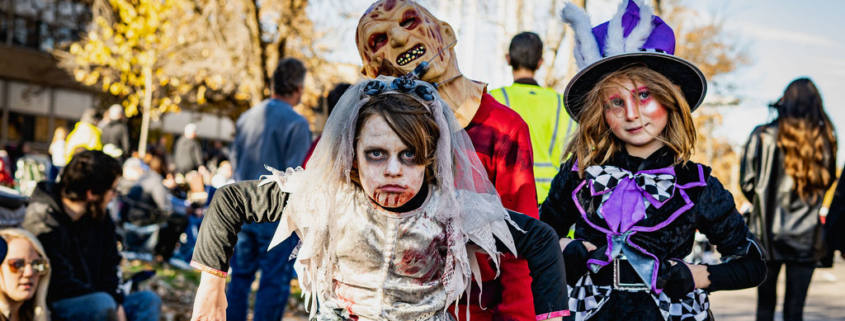 A young person in a bloody zombie bride costume looks directly at the camera, flanked by another person in a scary, monstrous mask and a person in a whimsical, Mad Hatter-style costume during a Halloween parade.