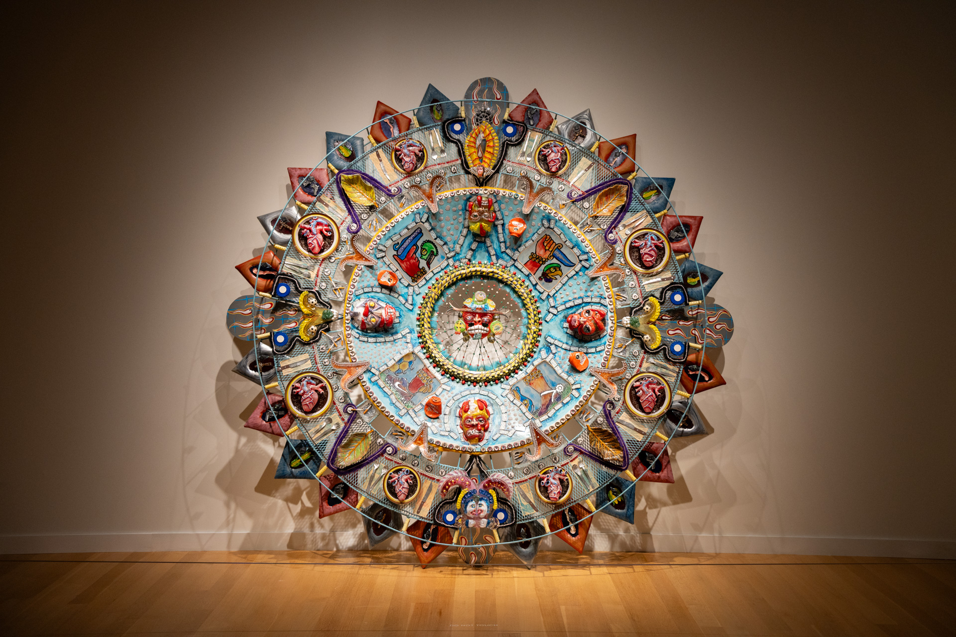 A head-on view of a large, colorful, and intricate circular sculpture, resembling a mandala, mounted on a beige wall in a museum.