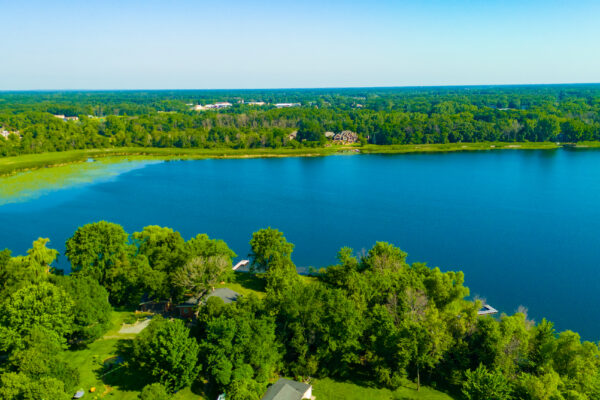 Aerial drone view of a large, deep blue lake surrounded by a dense green forest landscape and shoreline homes under a clear sky.