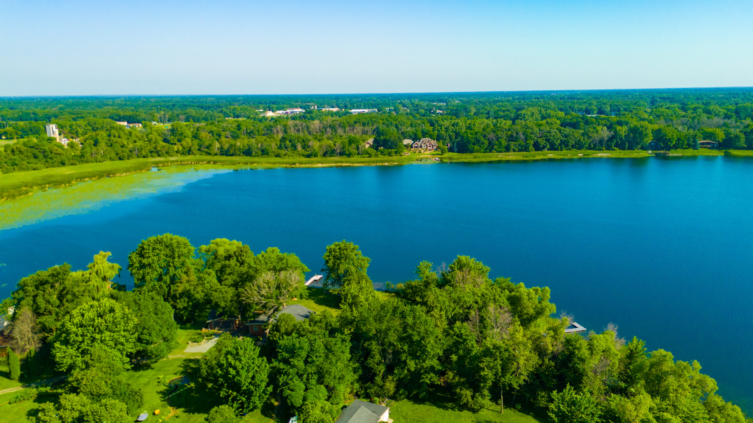 Aerial drone view of a large, deep blue lake surrounded by a dense green forest landscape and shoreline homes under a clear sky.