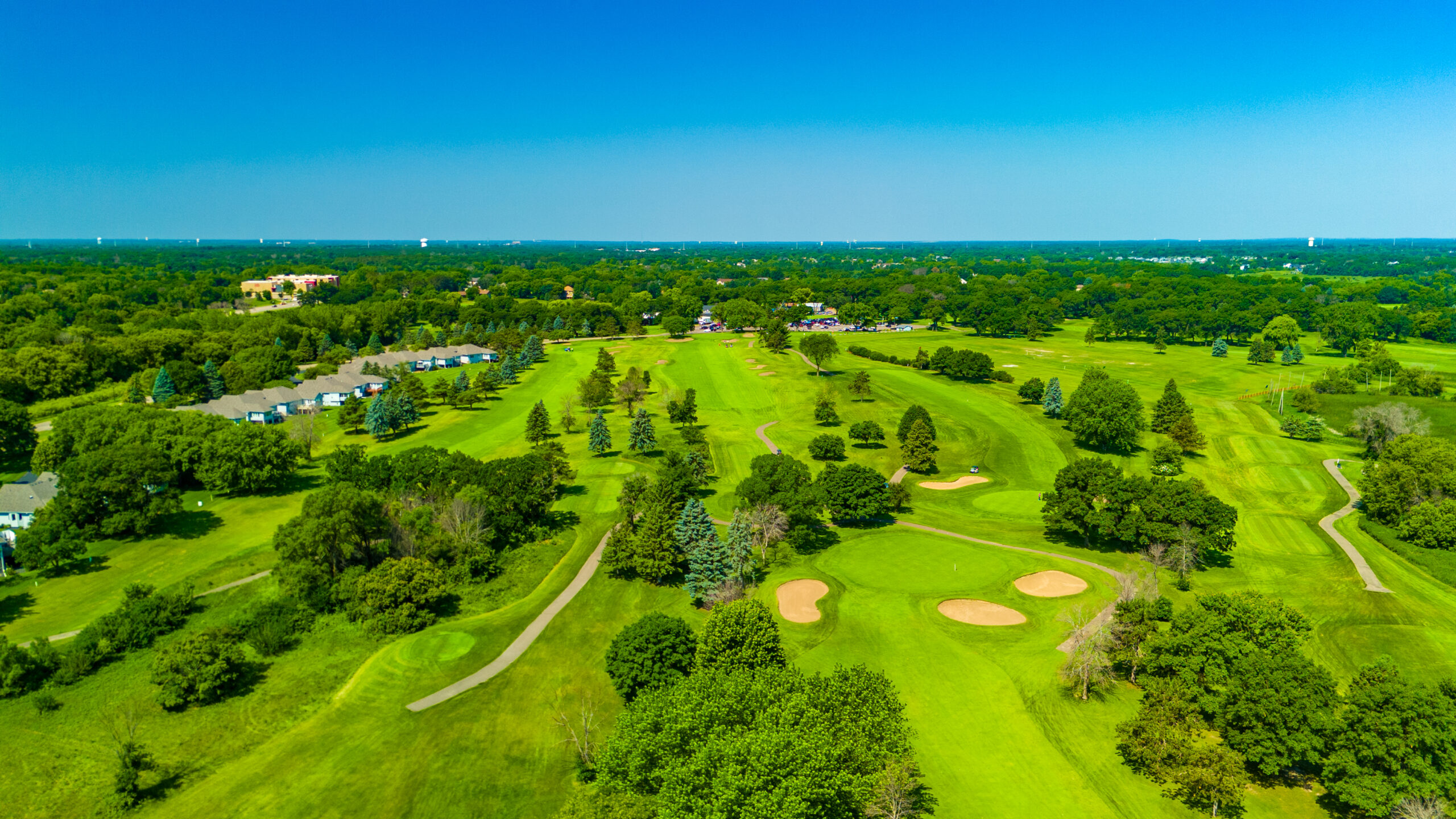 An aerial drone shot of the expansive, green fairways, sand traps, and mature trees of Majestic Oaks Golf Club under a clear blue sky.