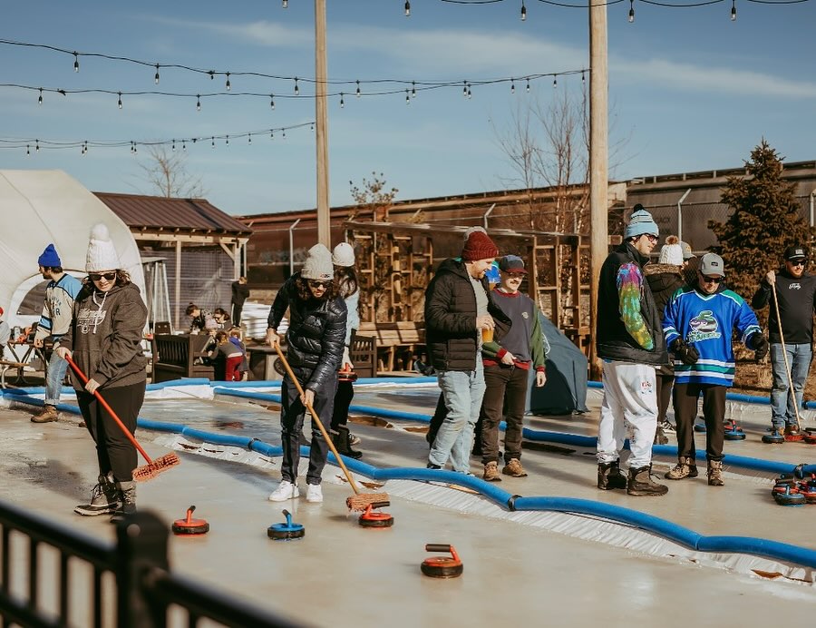 A group of young adults playing curling on a narrow outdoor ice rink bordered by blue bumpers. One person in a grey hoodie sweeps the ice with a red broom, while others stand nearby watching and socializing. The background features wooden structures and string lights against a clear blue sky.
