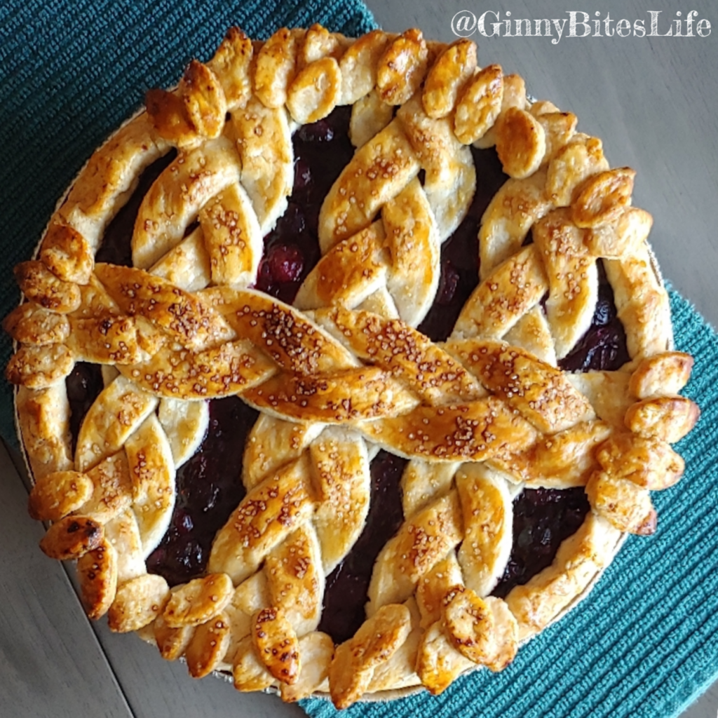 Top-down view of a golden-brown cherry pie from Ginny Bites with an intricate braided lattice crust and leaf-shaped dough accents around the edge.