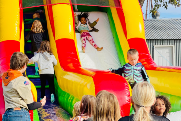 A group of children playing on a large, colorful inflatable slide. One young girl is sliding down the white center lane with her arms raised in excitement, while other children climb the blue stairs on the left. In the foreground, several children wait their turn at the base of the slide.