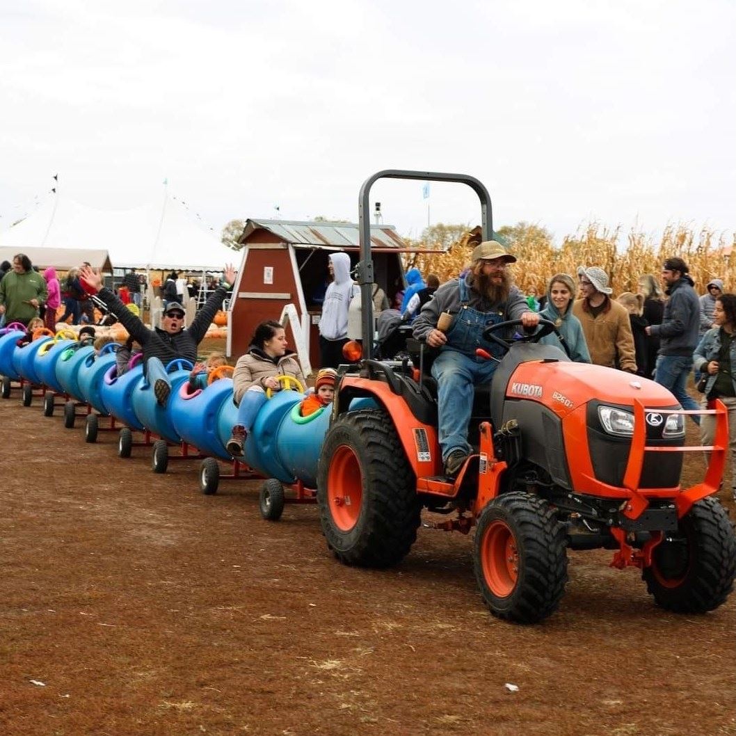 A line of children and adults riding in a blue barrel train being pulled by an orange Kubota tractor across a dirt field. The driver, a man with a beard and overalls, steers the tractor while passengers in the barrels wave their hands and smile. In the background, there are fair tents, a small red shed, and a crowd of people.