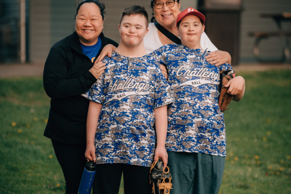 Two smiling boys with Down syndrome in matching "Challenger" baseball jerseys posing outdoors on the grass, one holding a glove, the other with a water bottle, with two supportive women standing behind them, smiling and with arms around their shoulders.